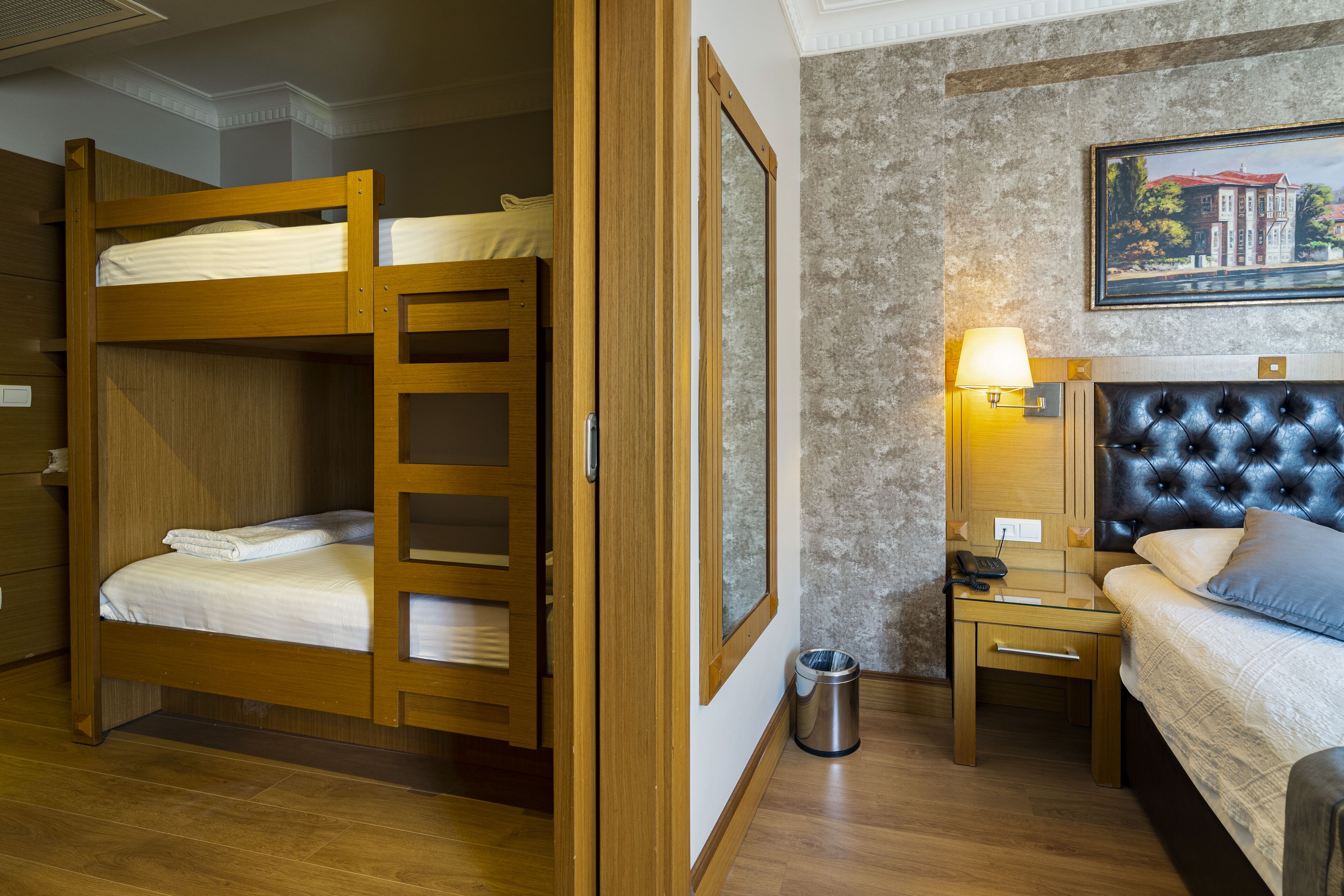 A hotel room with a bunk bed, a single bed, a bedside table with a lamp, a trash bin, and a framed picture on the wall.