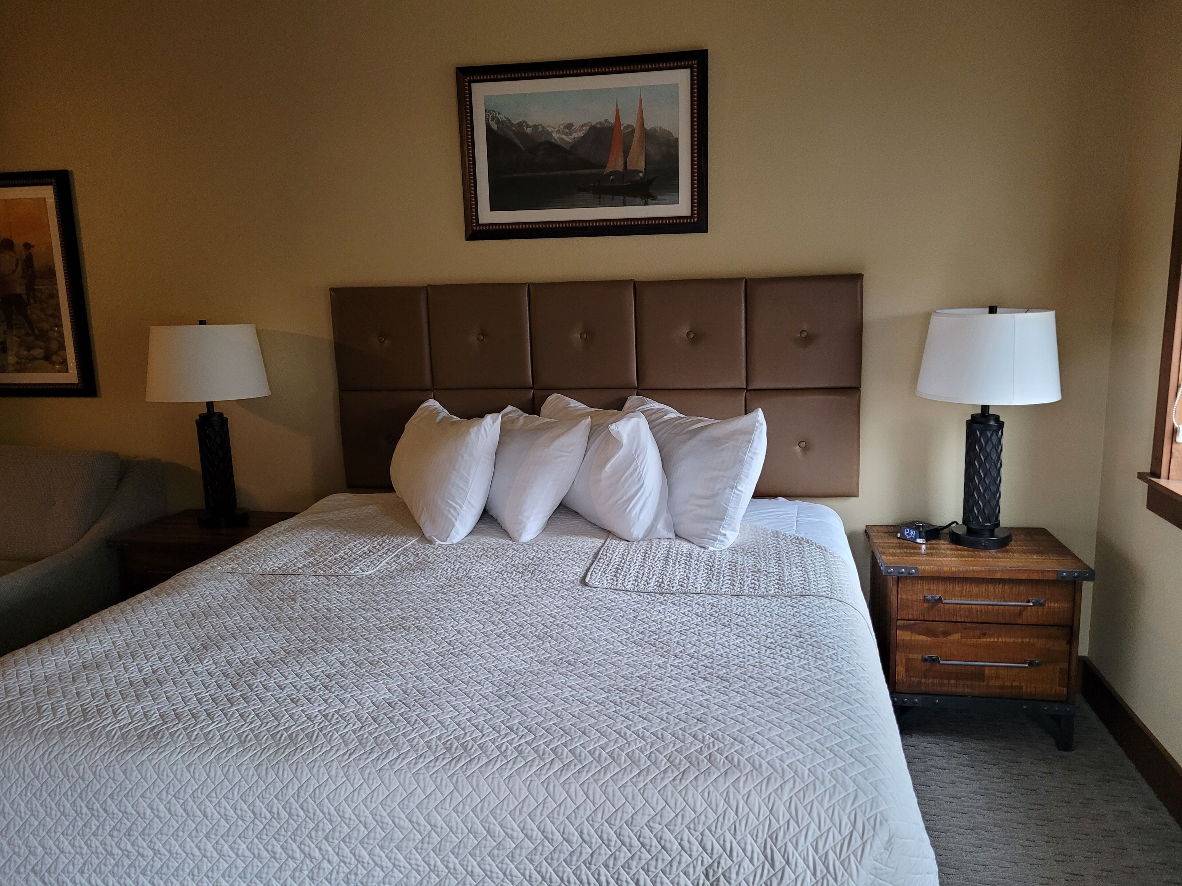 Una habitación de hotel con una cama grande, dos mesitas de noche con lámparas, un cabecero y un cuadro enmarcado en la pared.