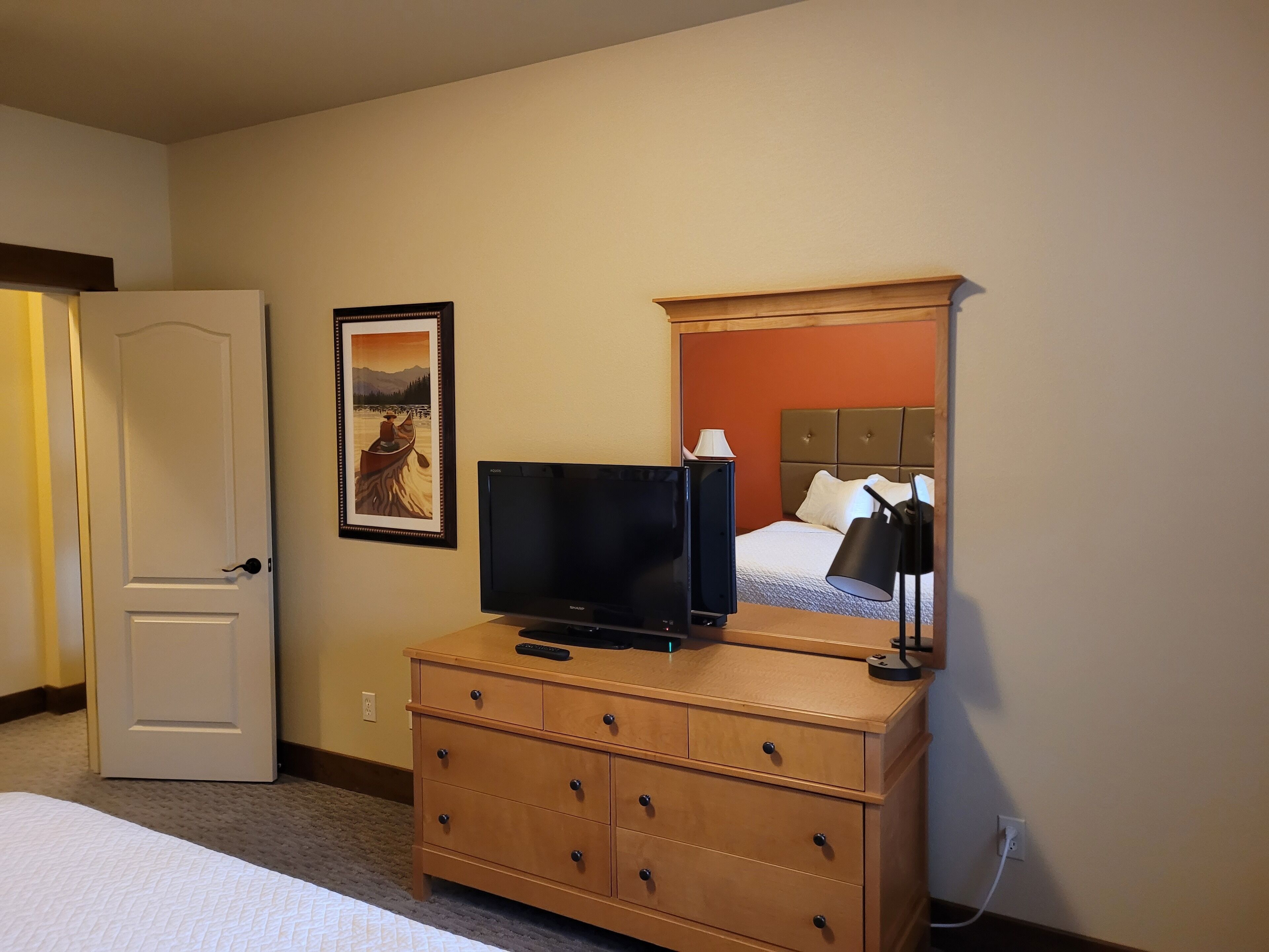 Habitación de hotel con cama, cómoda, televisor, un cuadro enmarco y espejo que refleja una cama y una lámpara.