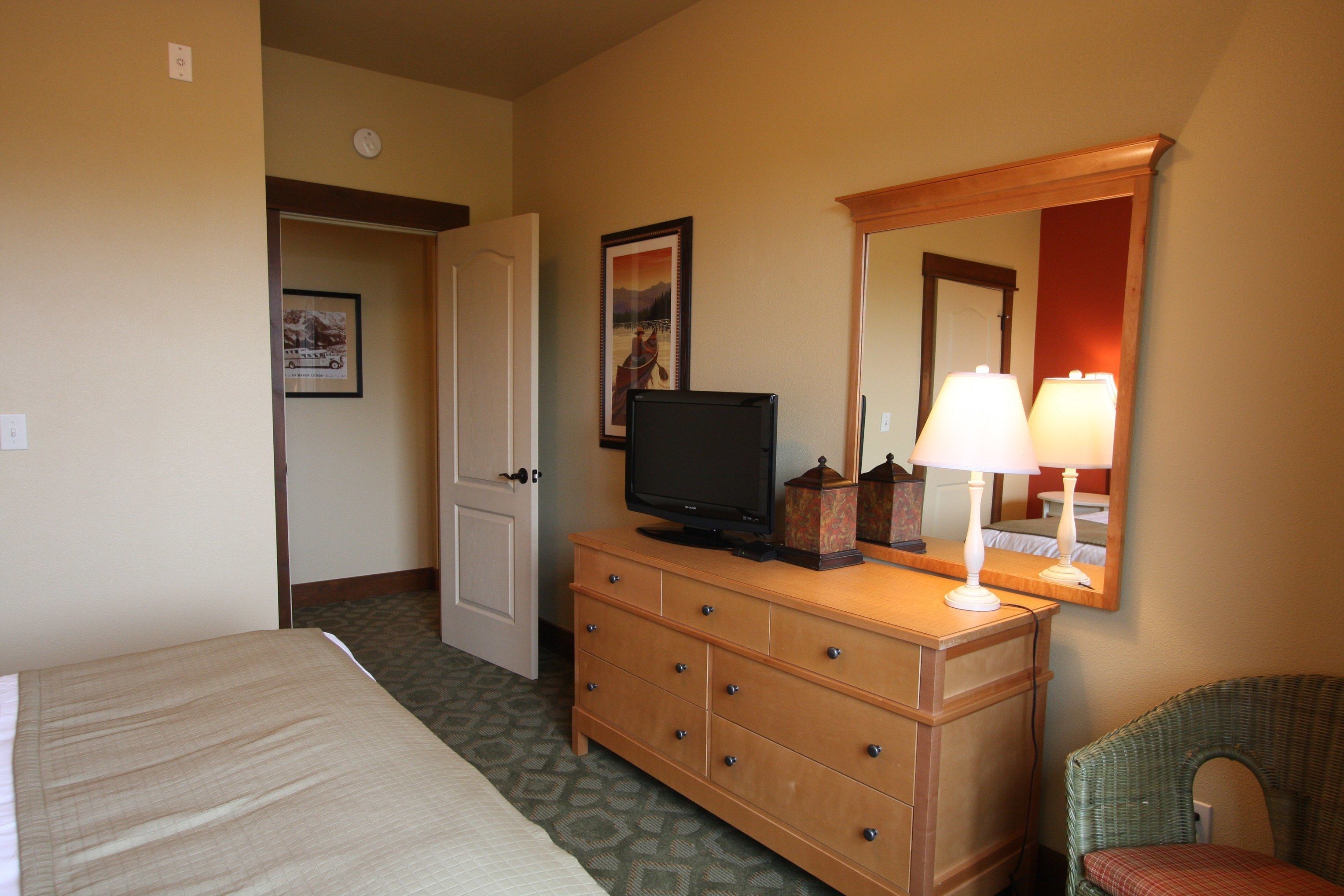 A hotel room with a bed, a wooden dresser with a television, a mirror, and a lamp.