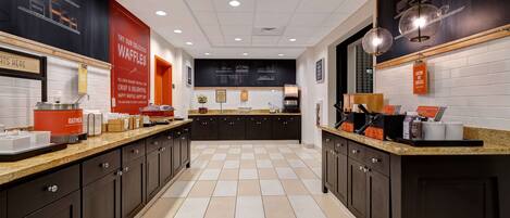 Uma cozinha moderna com armários escuros, piso ladrilhado e uma ilha central com diversos eletrodomésticos e utensílios.