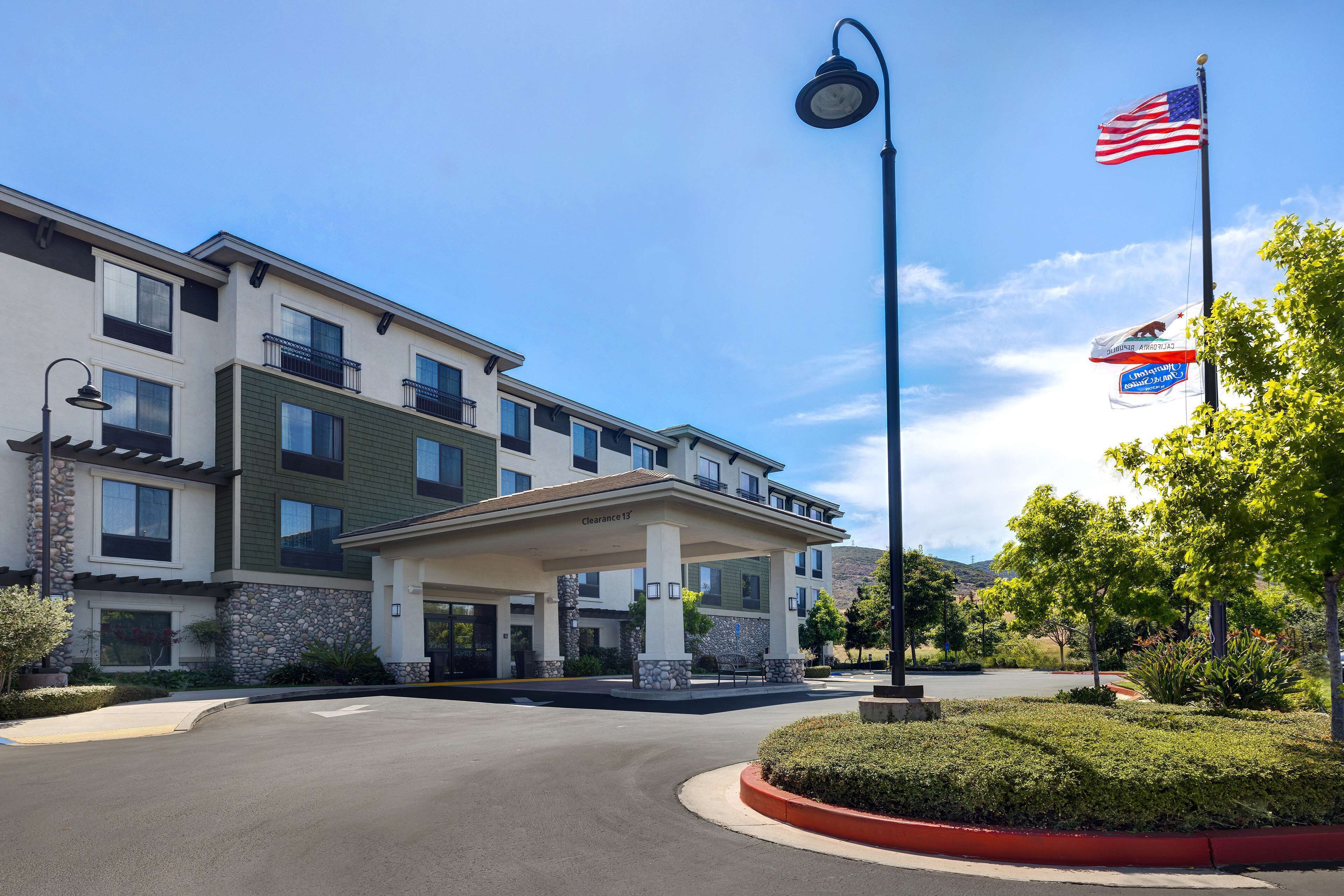 A multi-story hotel with a covered entrance, outdoor seating, and a landscaped area with a flagpole.