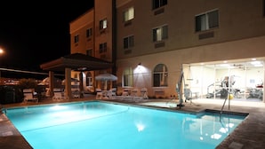 Una piscina con sillas de descanso y una zona cubierta, rodeada por un edificio con varias ventanas y un gimnasio visible a través del cristal.