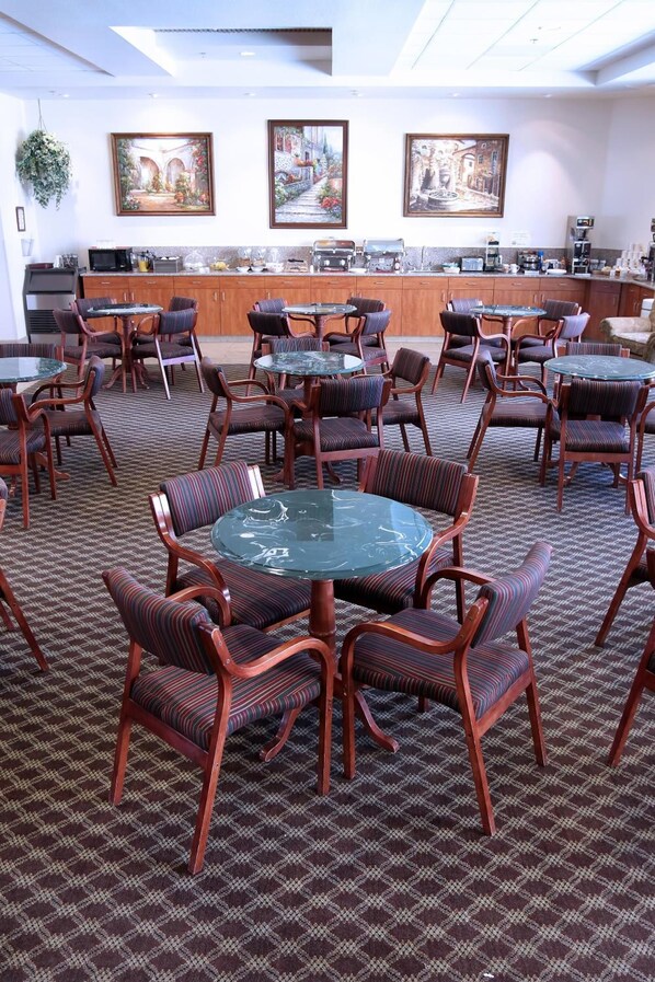 A conference room with round tables and chairs, a kitchen area, and three framed paintings on the wall. - Garden Place Suites (Sierra Vista)
