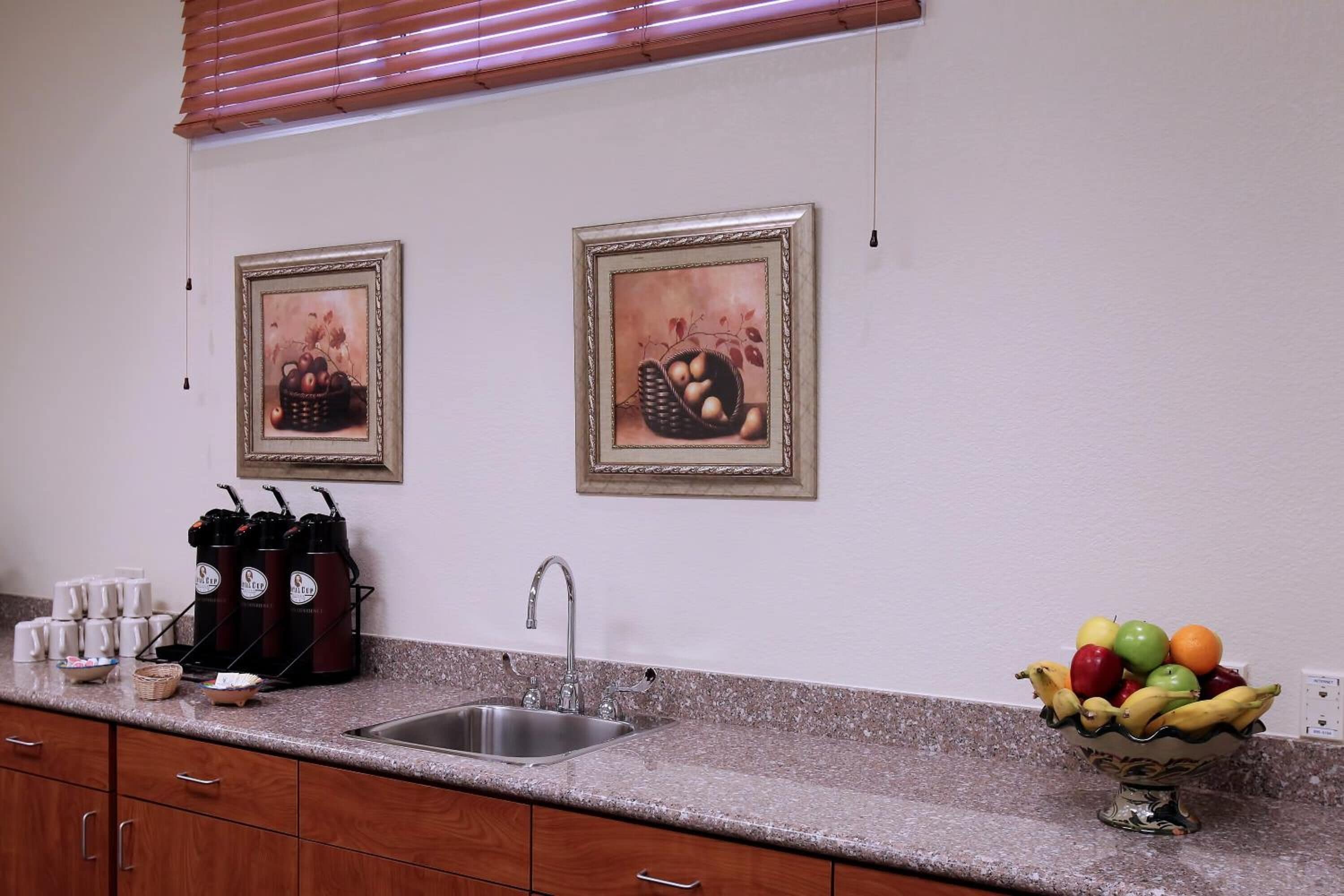 A kitchen with a granite countertop, a sink, and a fruit bowl. There are framed pictures on the wall and a coffee maker on the counter.