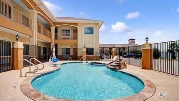 A swimming pool with a hot tub, surrounded by a fence, and a building with balconies in the background. - Crystal Inn Houston North I-45 (Houston)