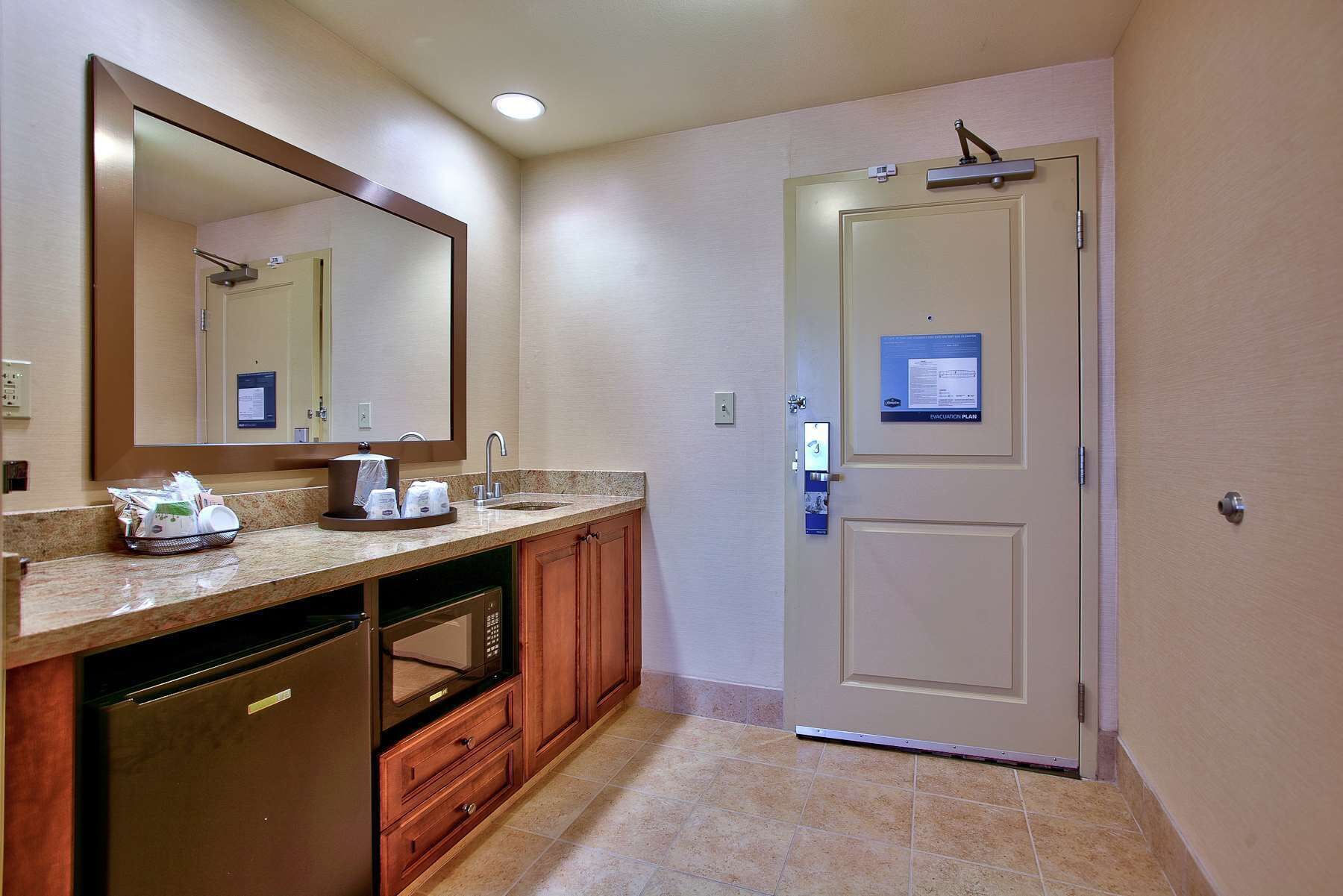A hotel bathroom with a sink, mirror, and a door with an emergency exit sign.
