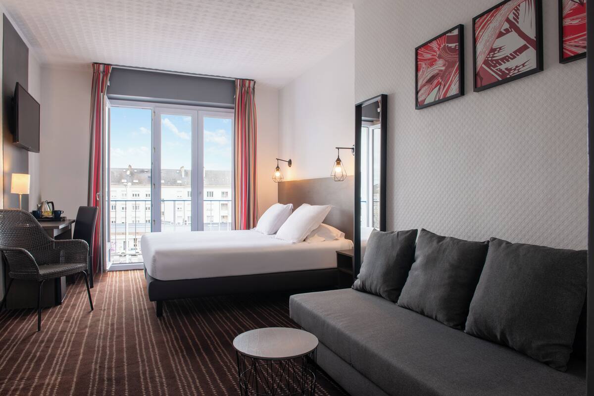 A hotel room with a bed, a sofa, a desk, and a chair. There is a window with curtains, a flat-screen TV, and wall-mounted lights.