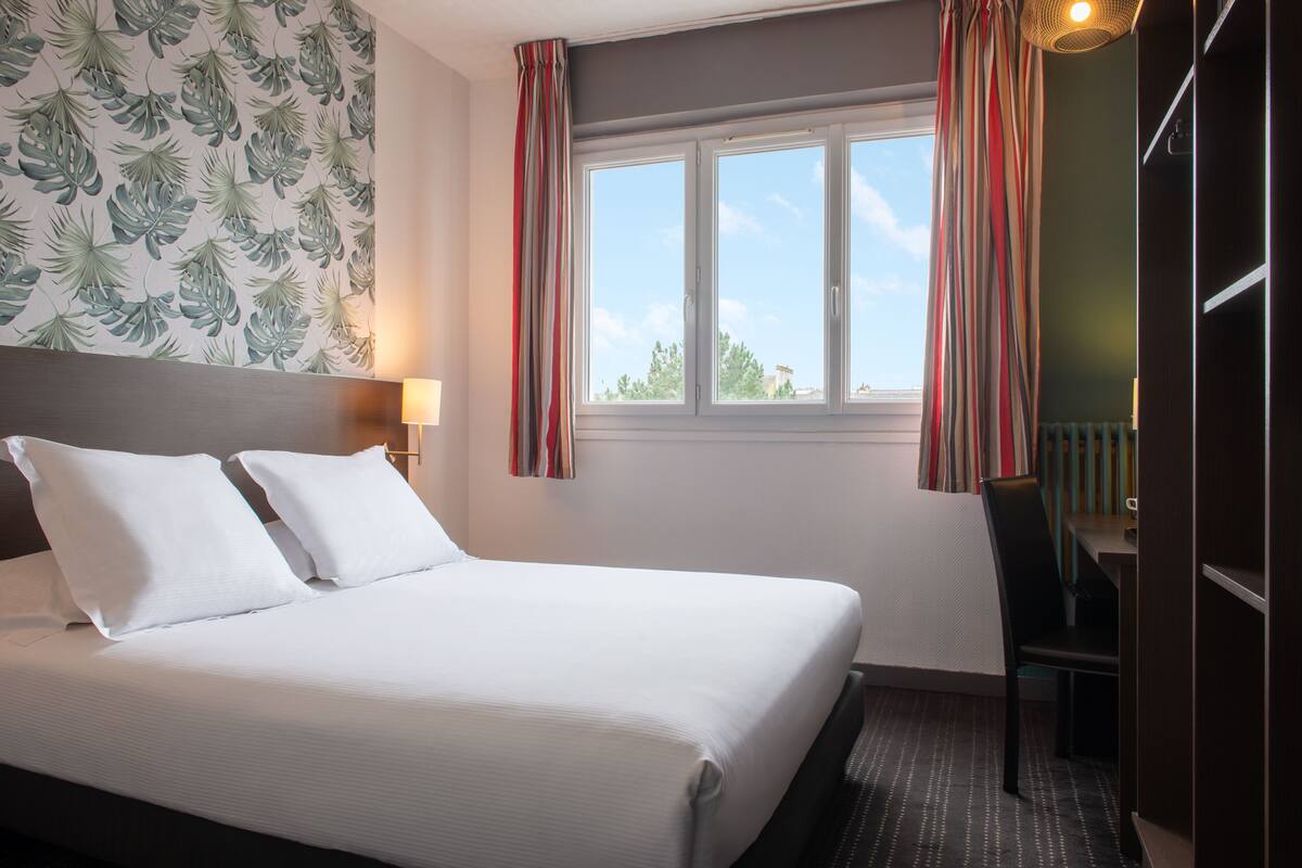 A neatly made bed with white linens, a window with curtains, a desk with a chair, and a wall with a leaf pattern.