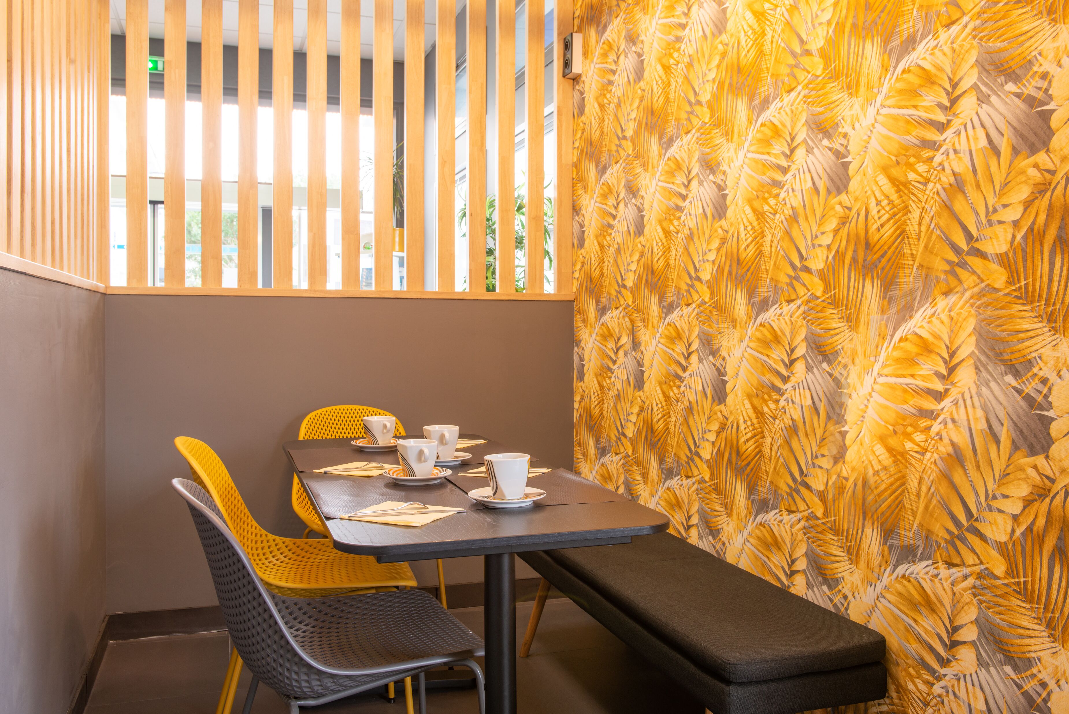 A dining area with a table set for two, featuring yellow and gray chairs, a bench, and a patterned wall.
