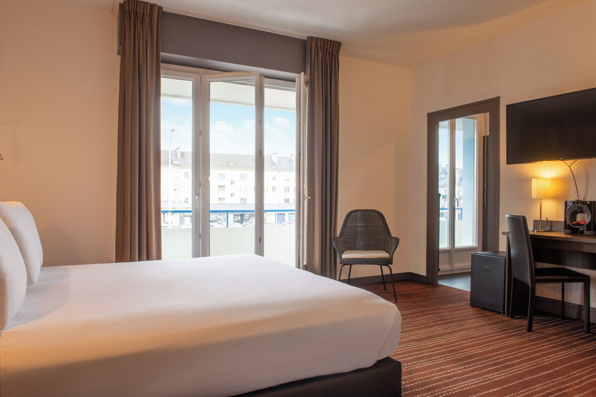 A hotel room with a large bed, a desk with a chair, a television, and a view of buildings through the window.
