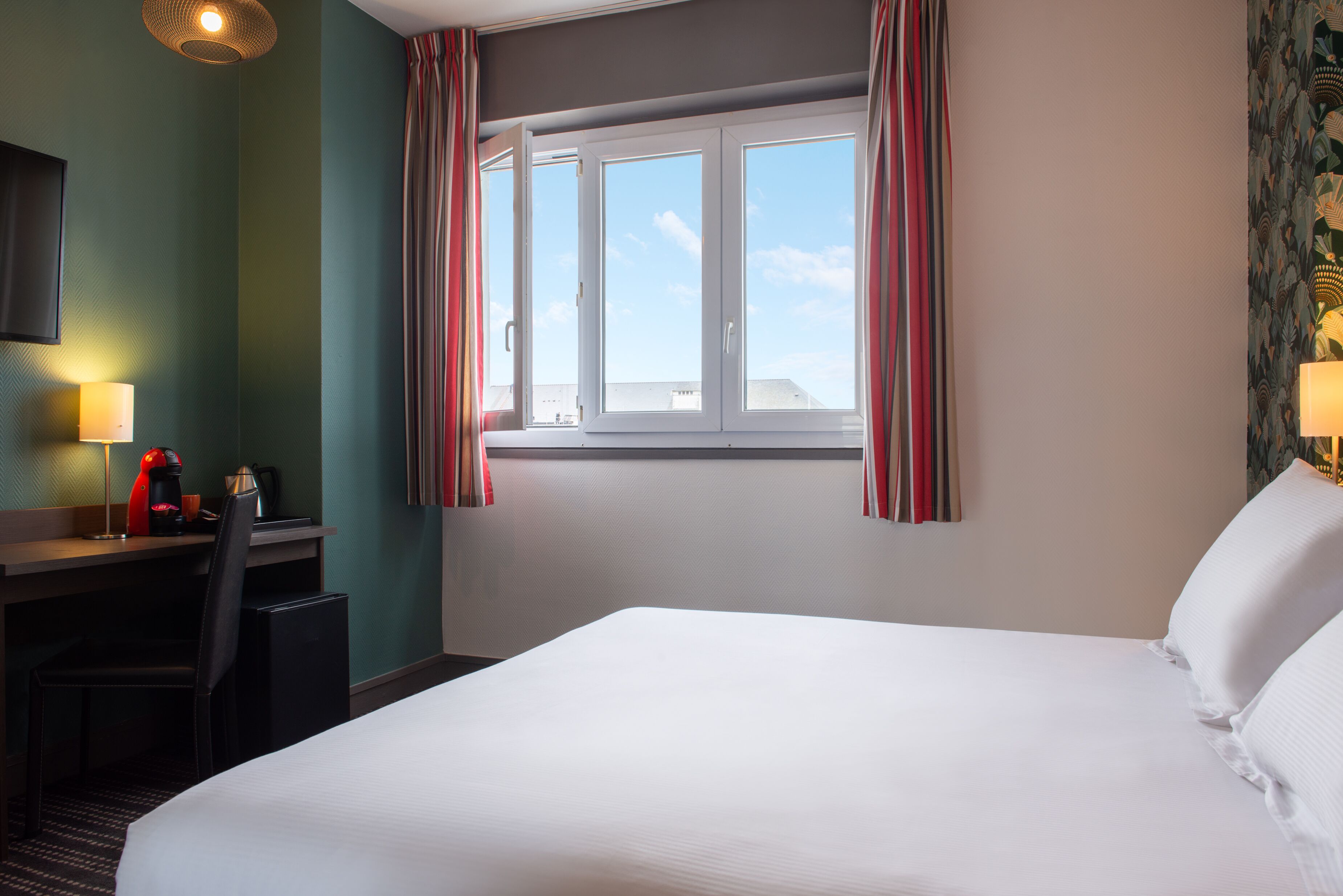 Habitación de hotel con una cama grande, un escritorio con silla, una cafetera y una ventana con cortinas.