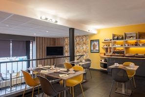 A modern restaurant with a mix of yellow and brown chairs, a flat-screen TV, and a variety of food items on display.