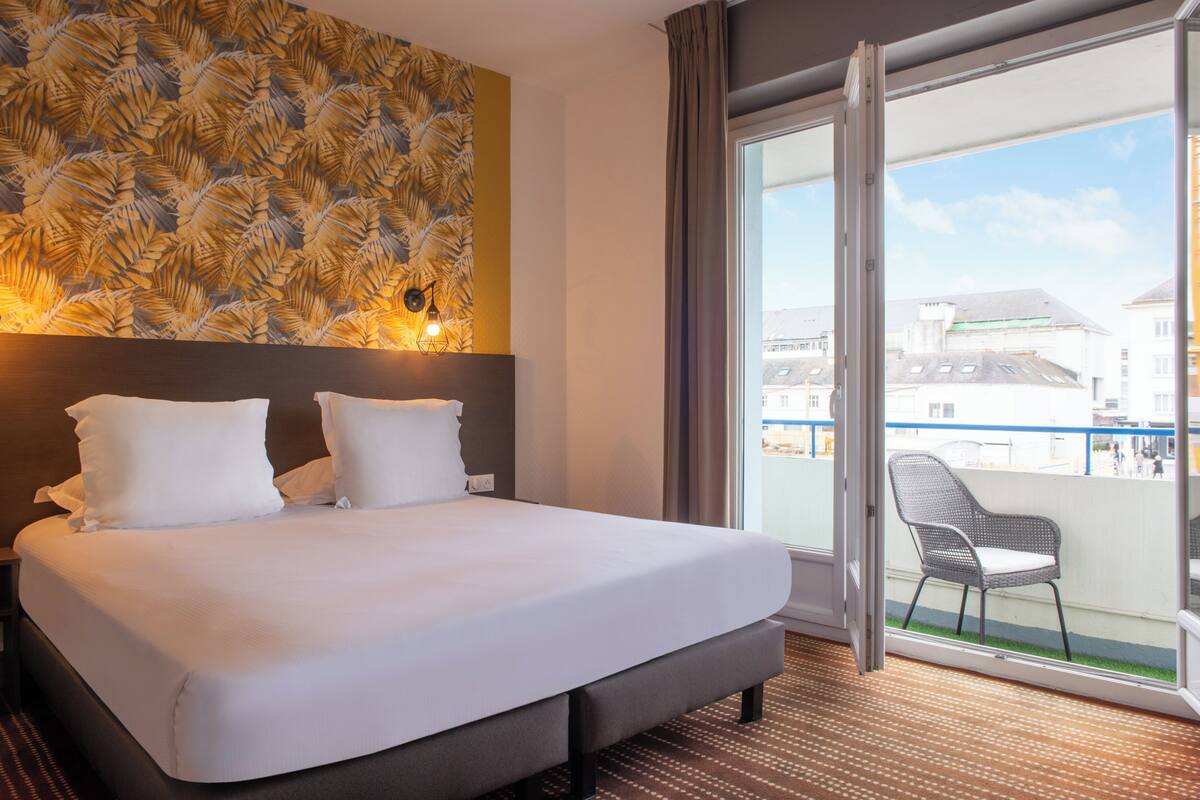 A hotel room with a large bed, a headboard with a leaf pattern, a balcony with a chair, and a view of buildings outside.
