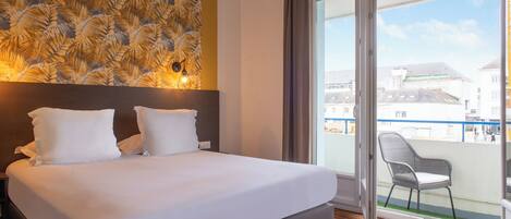 Quarto de hotel com uma cama grande, cabeceira com padrão de folha, varanda com uma cadeira e vista para edifícios.