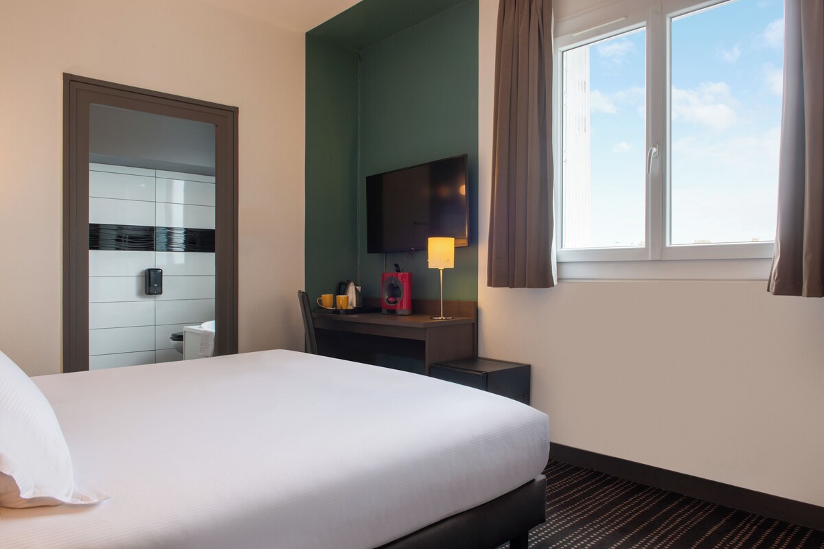 Una habitación de hotel con una cama, un escritorio con lámpara, un televisor y un ventanal con cortinas.