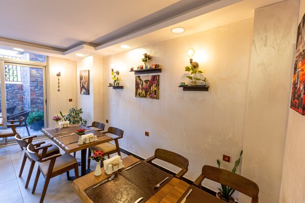Un comedor con mesas y sillas de madera, estantes fijos en la pared con objetos decorativos, y vista al exterior a través de un vidrio.