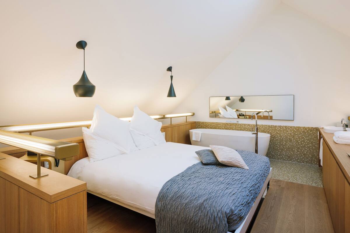 A modern hotel room with a bed, bedside table, lamp, and a bathroom with a sink and mirror.