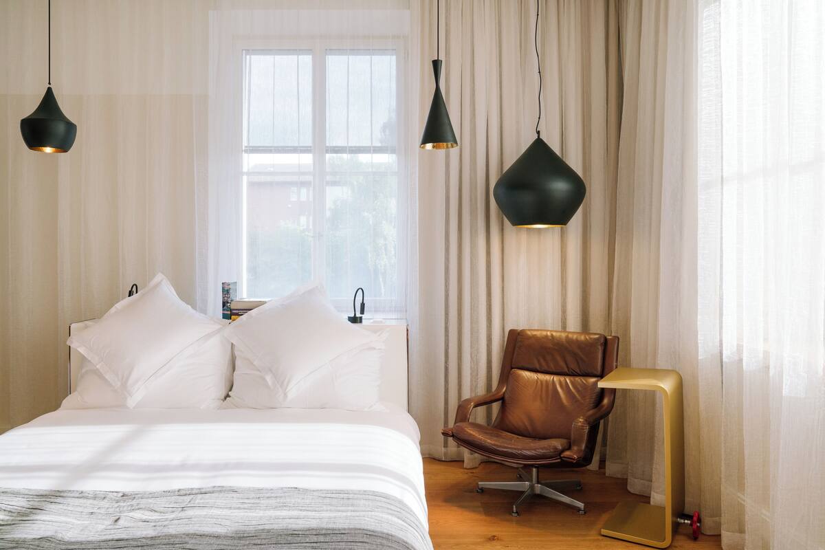 A bedroom with a bed, two hanging pendant lights, a leather chair, and a floor lamp.