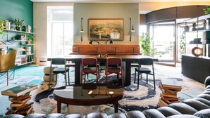 A modern living room with a round coffee table, two armchairs, and a sofa. There is a bookshelf filled with books and decorative items. A painting hangs on the wall above the coffee table. - B2 Hotel Zürich (Zürich)