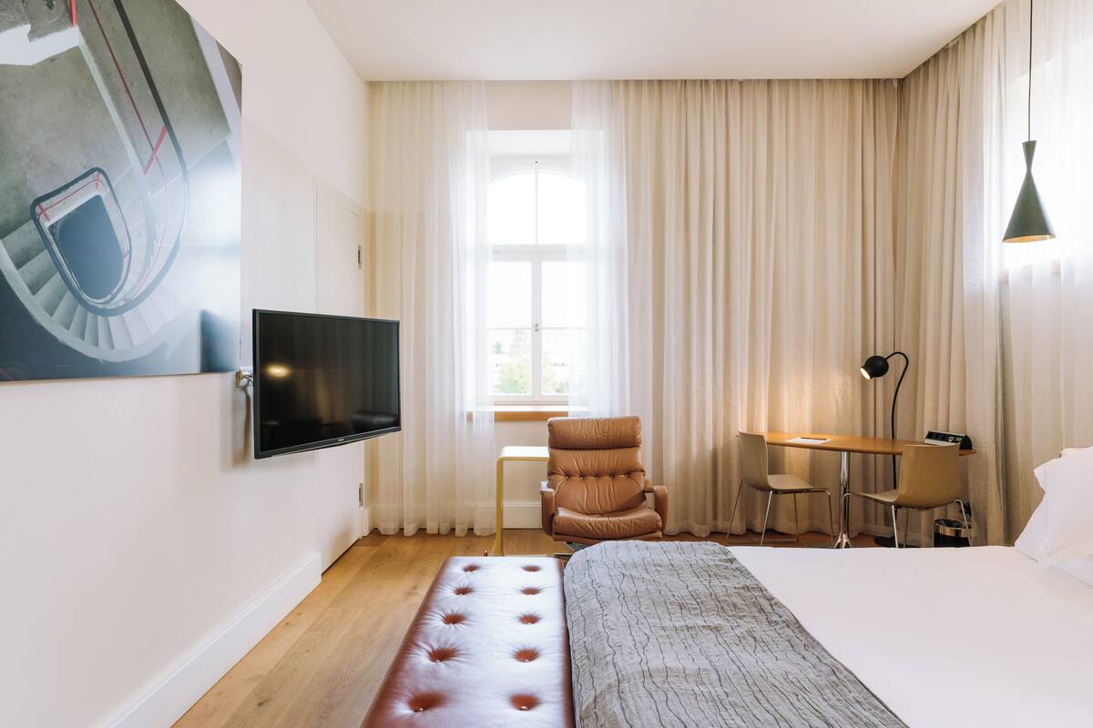 A hotel room with a bed, a desk, a chair, a TV, and a window with curtains.