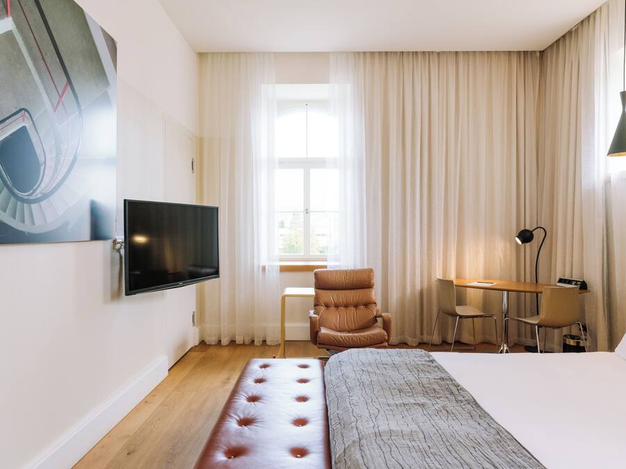 Habitació d'hotel amb llit, escriptori, cadira, televisió i finestra amb cortines.