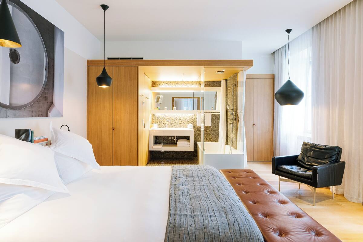 A modern hotel room with a large bed, a sitting area, a bathroom with a mirror, and a view of the outside.