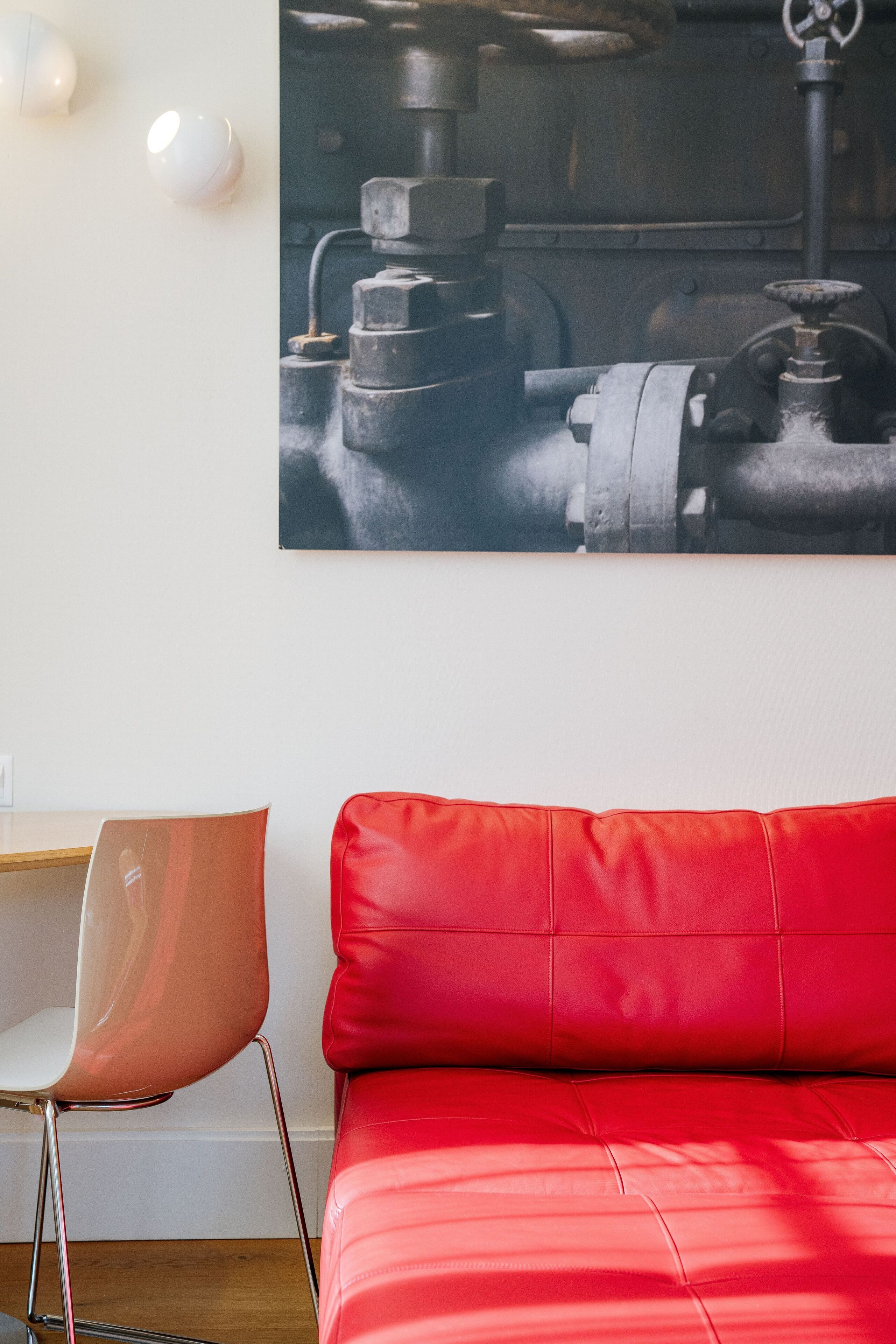 Una habitación moderna con un sofá de cuero rojo, un escritorio de madera y una silla. Un cuadro de gran formato con temática industrial adorna la pared.