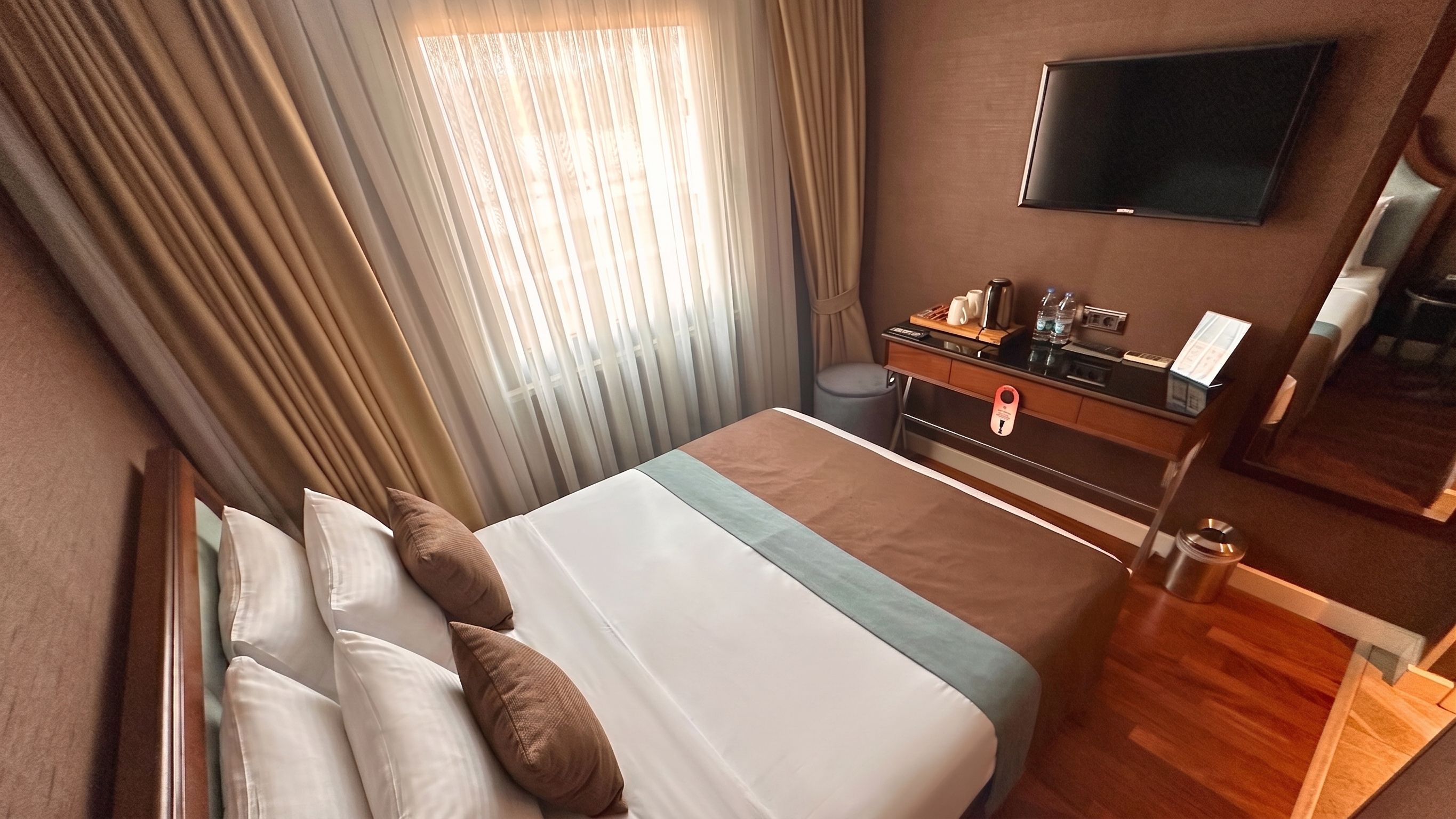 Une chambre d’hôtel avec un lit, un bureau doté d’un téléviseur à écran plat, une fenêtre avec des rideaux et un plancher en bois.