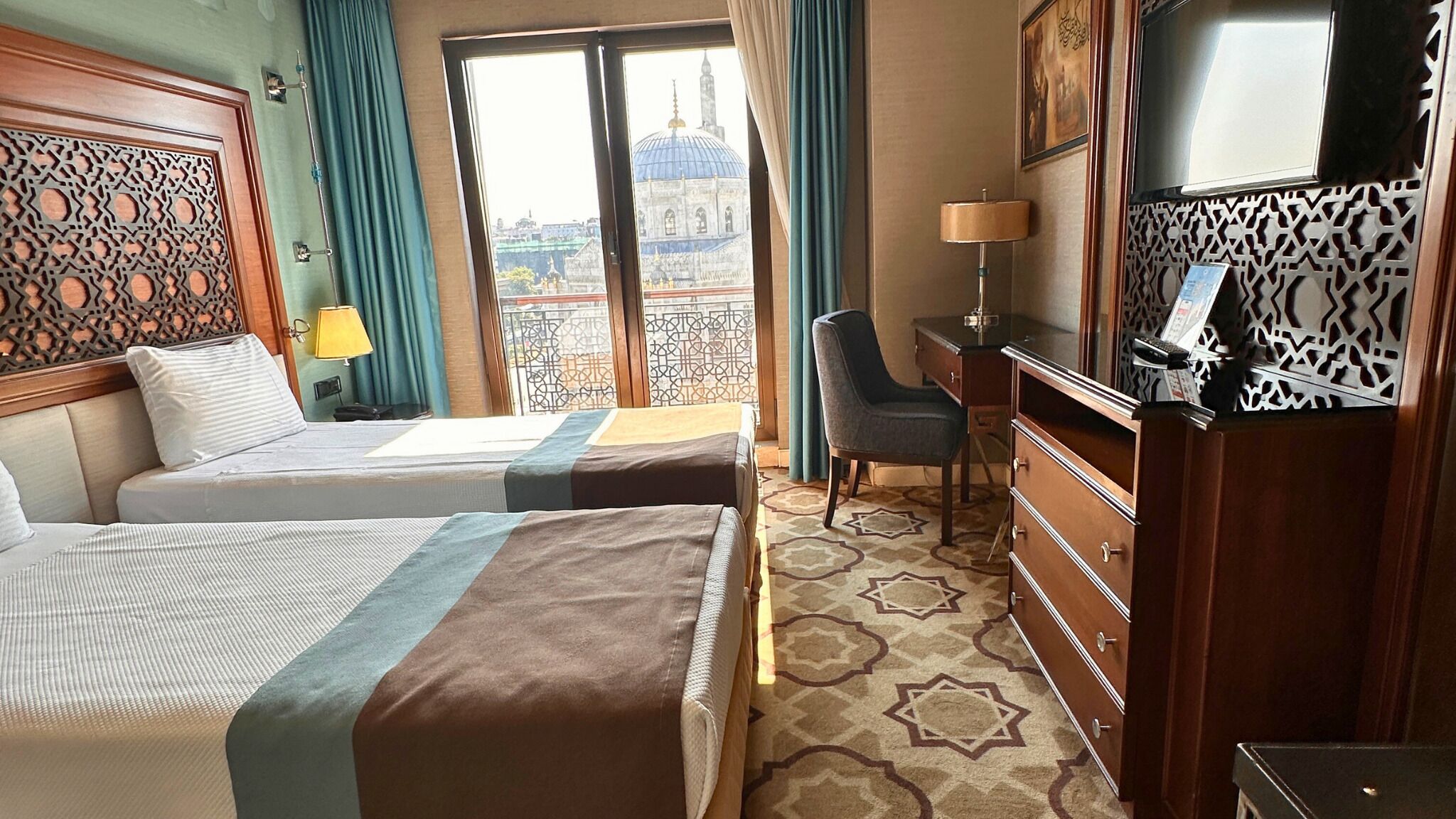 Une chambre d’hôtel avec deux lits, un bureau, une chaise, une télévision et une vue sur une mosquée par la fenêtre.