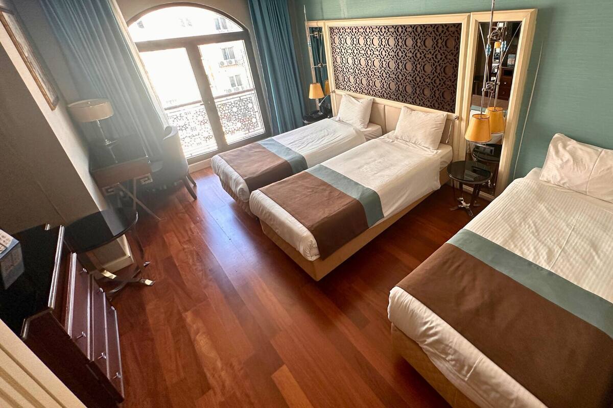 A hotel room with two beds, a wooden floor, a decorative headboard, a small table with a lamp, and a view of the outside.