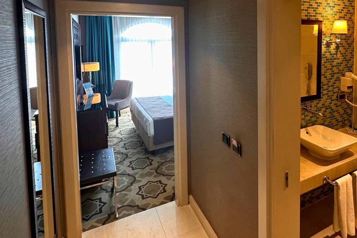 A hotel room with a bed, bedside table, lamp, and a view of a hotel lobby.