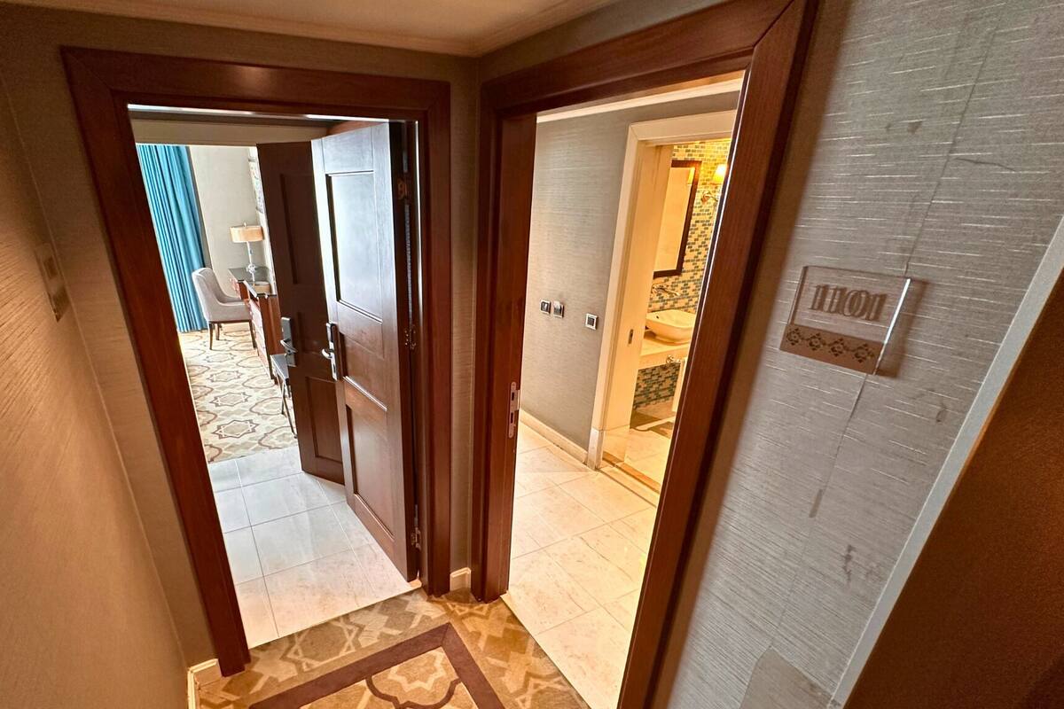 A hallway with a patterned carpet, a wooden door, and a bathroom with a tiled wall and a sink.