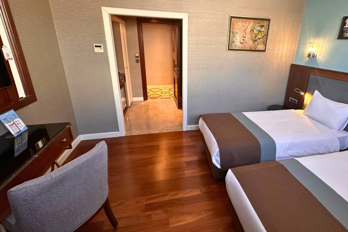 A hotel room with two beds, a desk with a chair, a small table, and a door leading to a hallway.