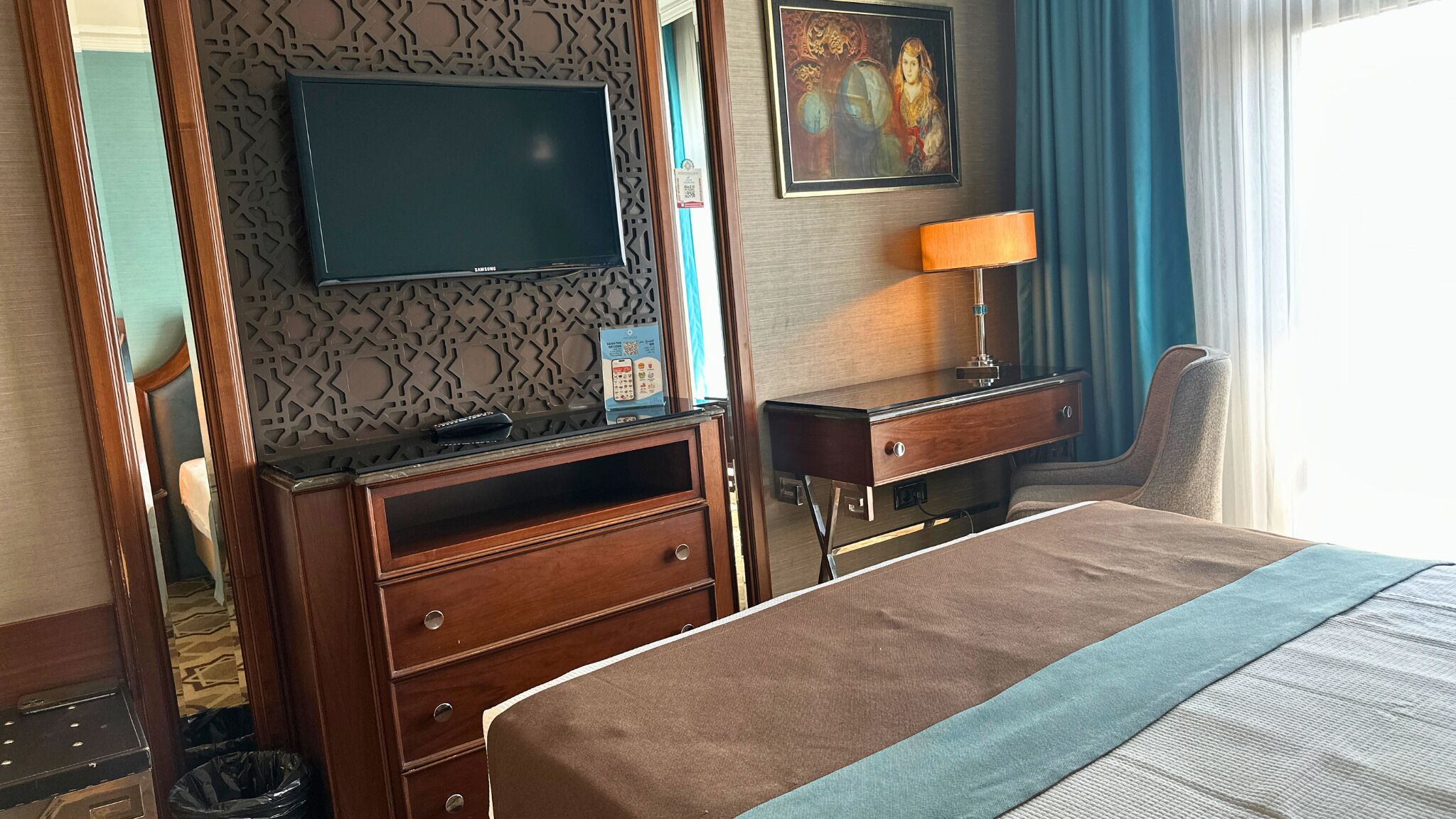 A hotel room with a bed, a desk with a lamp, a television, and a painting on the wall.