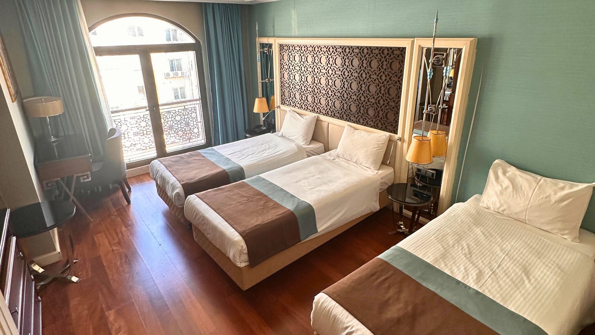 A hotel room with two beds, a wooden floor, a patterned headboard, a small table with a lamp, and a view of the outside balcony.