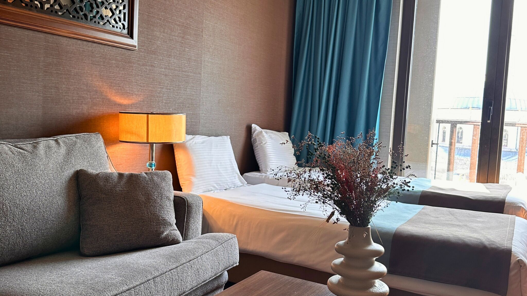 A hotel room with a sofa, a bed, a nightstand, a lamp, and a window with curtains.