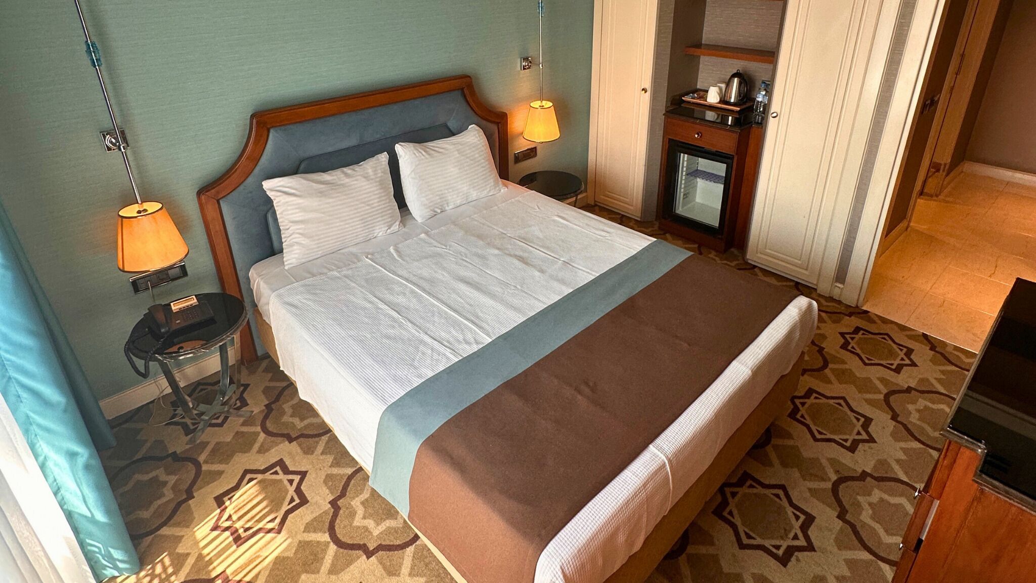 A hotel room with a large bed, two bedside lamps, a small table with a telephone, and a minibar.
