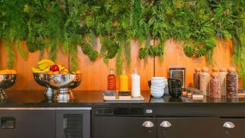 Disposição de buffet com diversos alimentos e acompanhamentos, um espaço para café e uma parede decorativa com plantas penduradas.