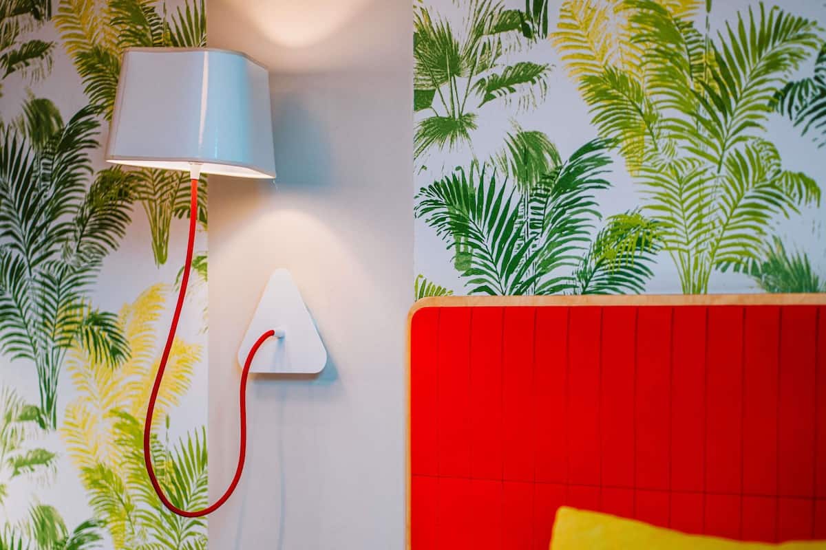 A room with a red headboard, a white wall with a leaf pattern, a white lamp, and a power outlet.