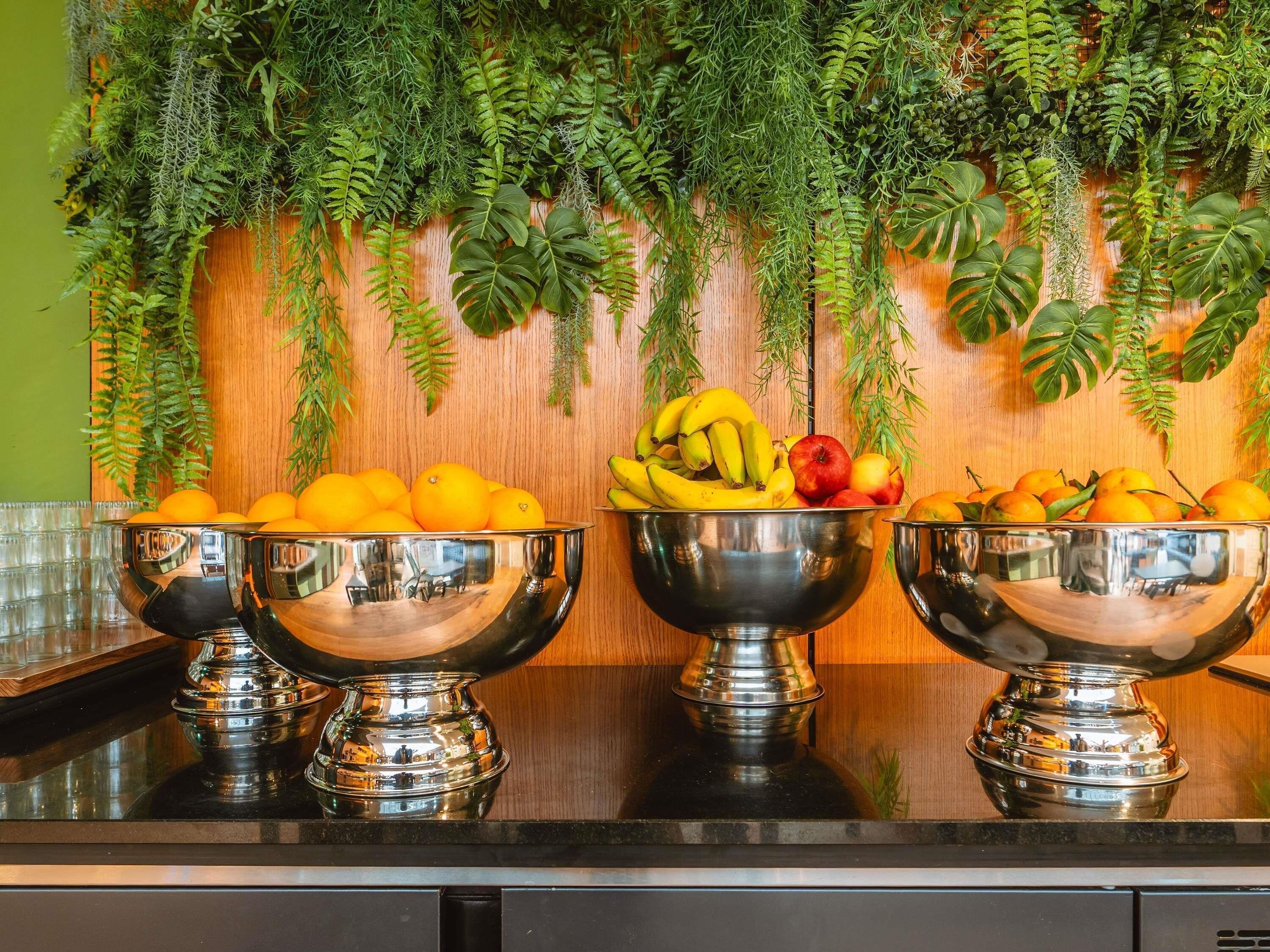 A kitchen counter with three silver bowls containing fruits, a wooden wall with hanging plants, and a green wall in the background.
