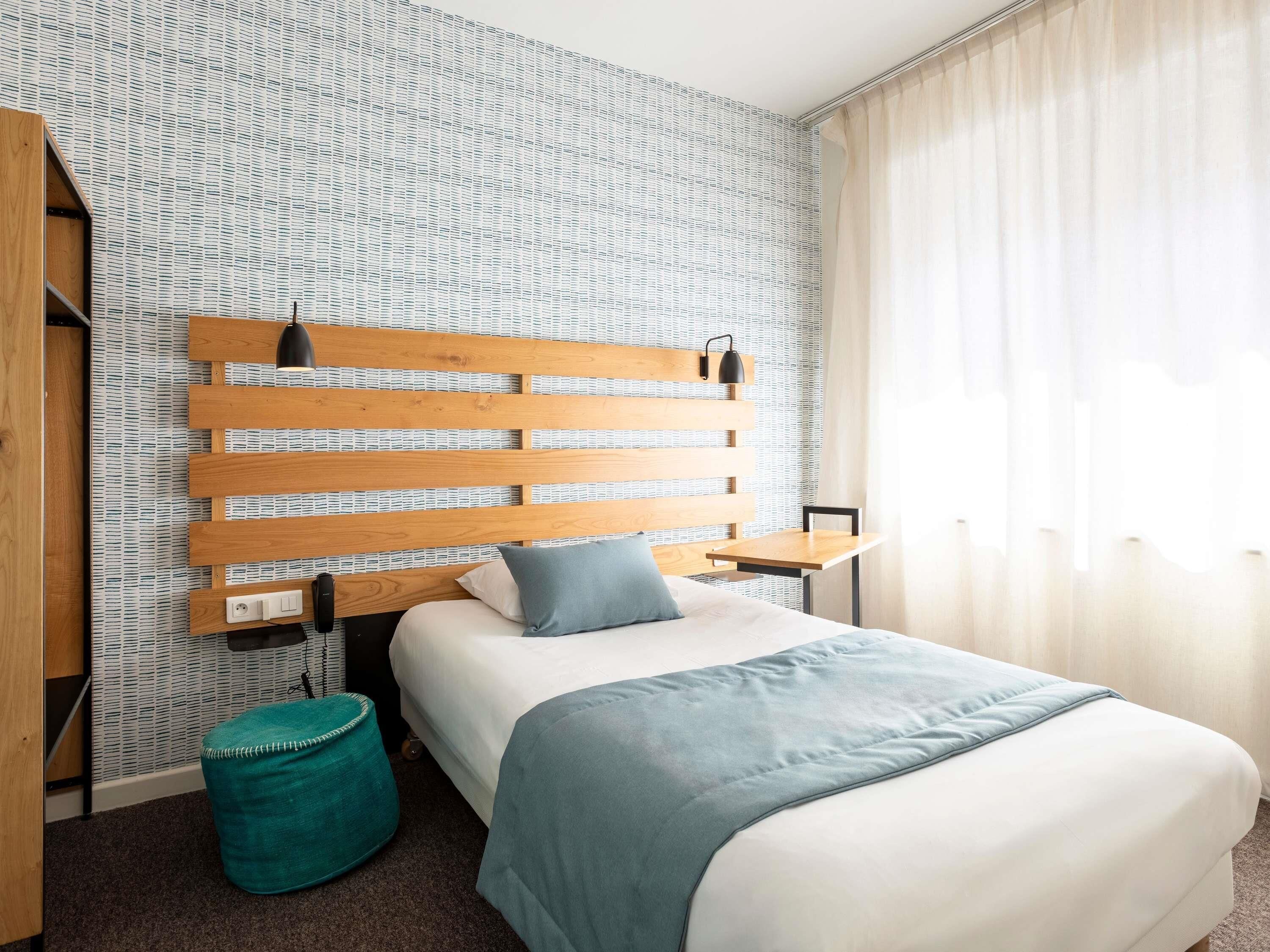 Une chambre d’hôtel avec un lit, une tête de lit en bois, une petite table, un pouf turquoise et une fenêtre avec des rideaux.