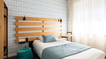 A hotel room with a bed, a wooden headboard, a small table, a teal ottoman, and a window with curtains.