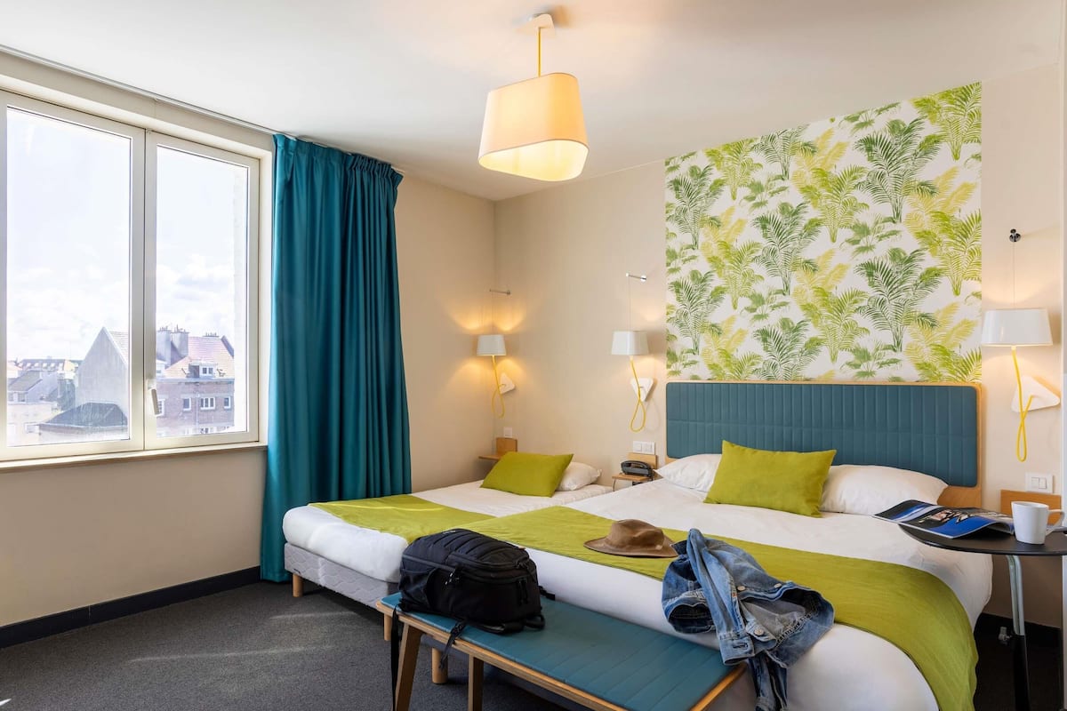 A hotel room with a bed, bedside tables, a bench, a window with curtains, and a wall with a leaf pattern.