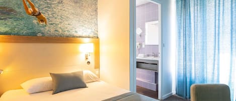 Habitación de hotel con cama, mesita de noche, lámpara de pared, baño visible a través de una puerta abierta y una silla.