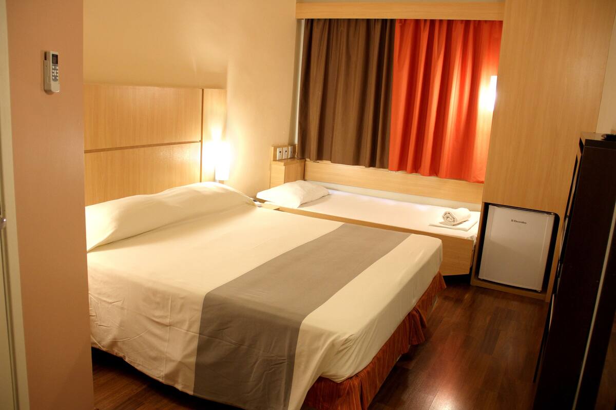 Habitación de hotel con cama, mesita de noche y un refrigerador pequeño.
