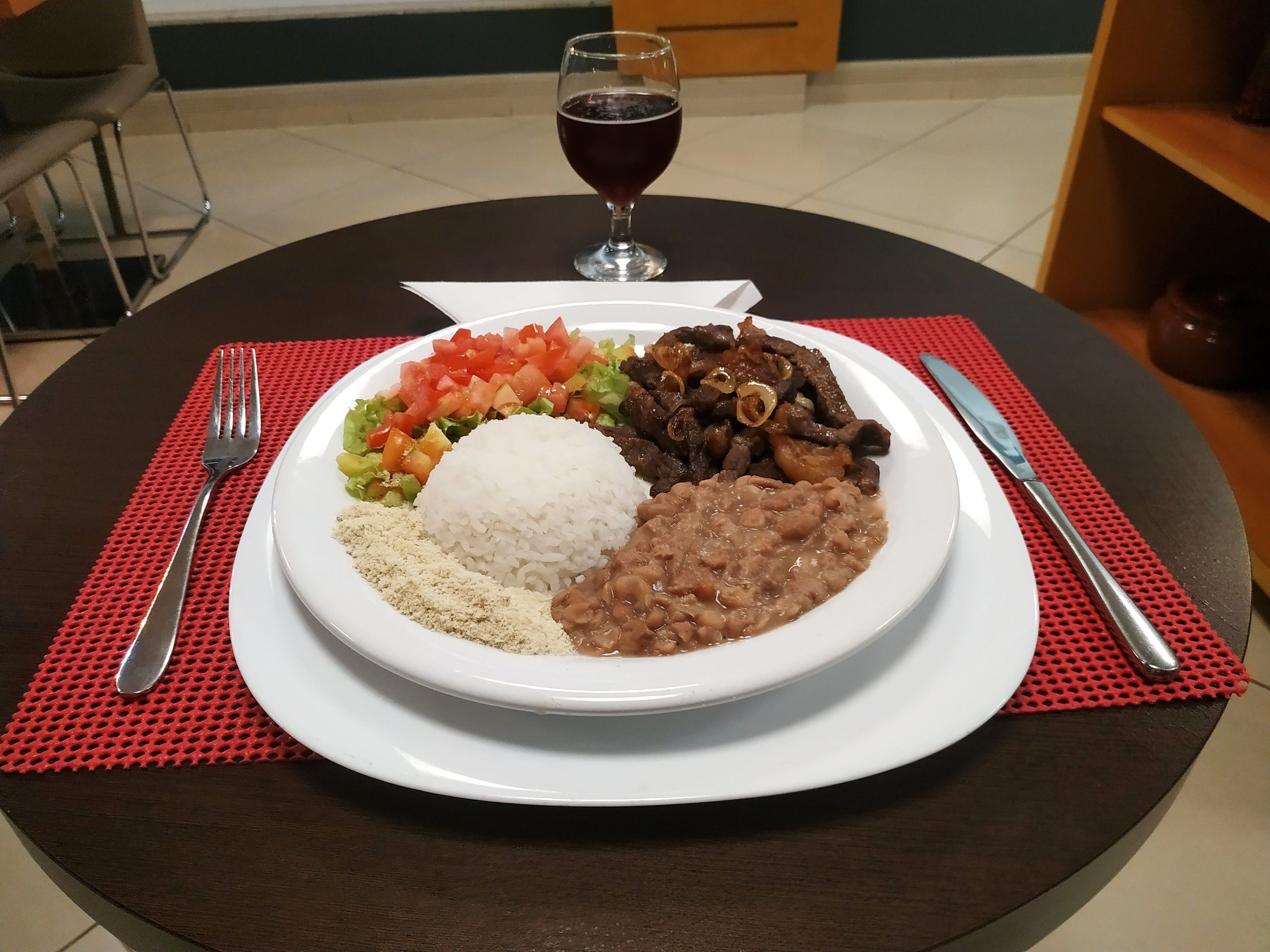 A plate of food with rice, beans, meat, and vegetables, accompanied by a glass of red wine on a table with a red placemat.