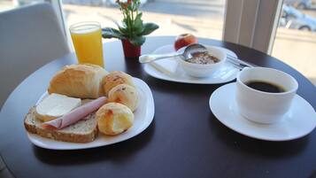 Una colazione con croissant, prosciutto e pane su un piatto, una ciotola di cereali, un bicchiere di succo d'arancia e una tazza di caffè su un tavolo.