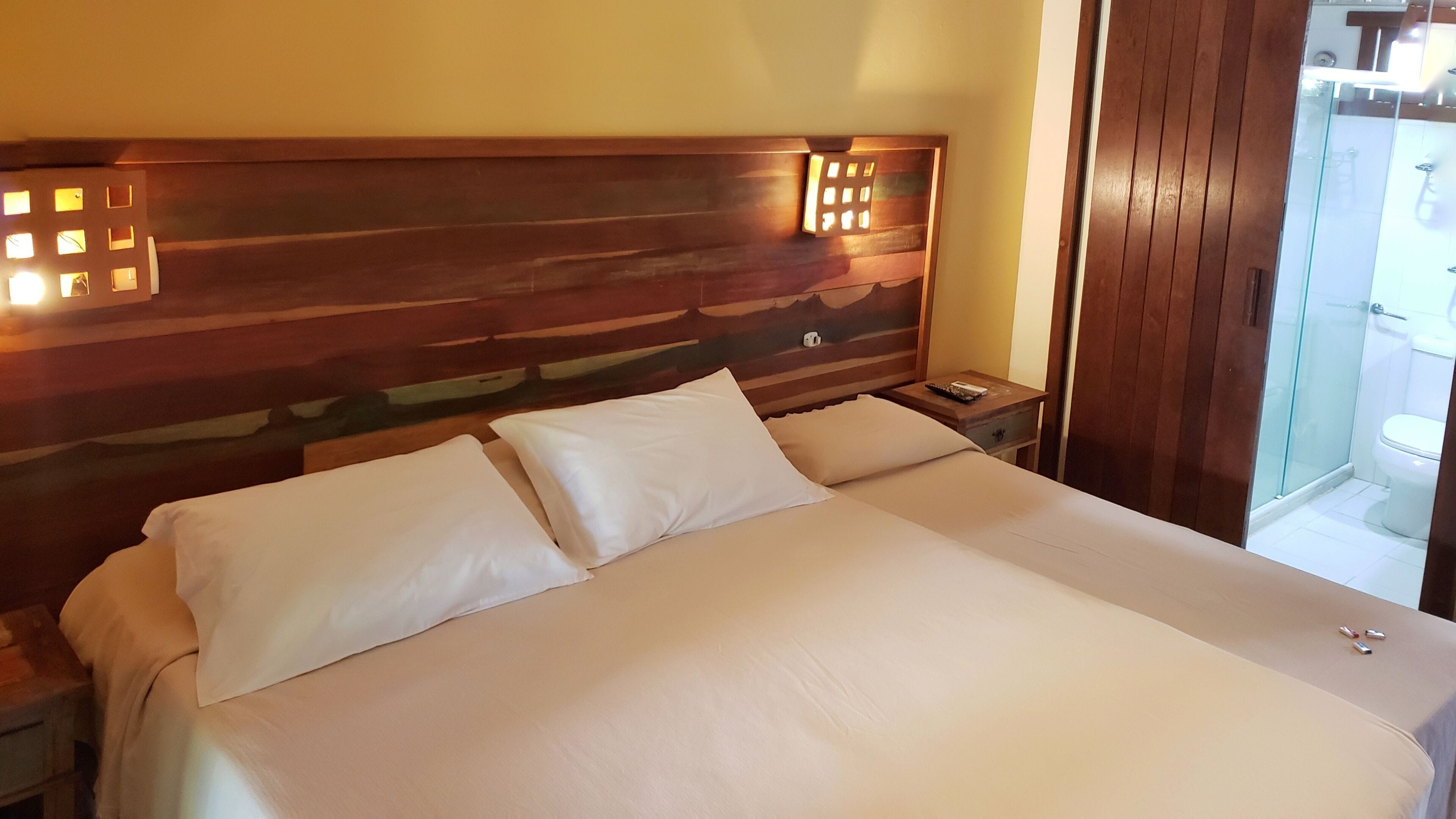 Une chambre d’hôtel avec un grand lit, une tête de lit en bois, des tables de chevet et une salle de bain visible à travers une porte en verre.