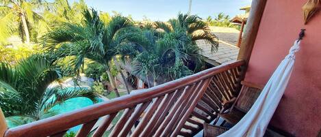 Un balcón con baranda de madera, una hamaca y vista a una piscina y palmeras.